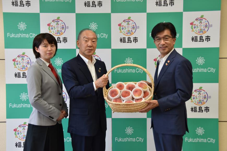 福島市のバックボードの前で、たくさんの桃が入ったかごを市長と女性と並んで立ったスーツの男性が左右から持って写った記念写真