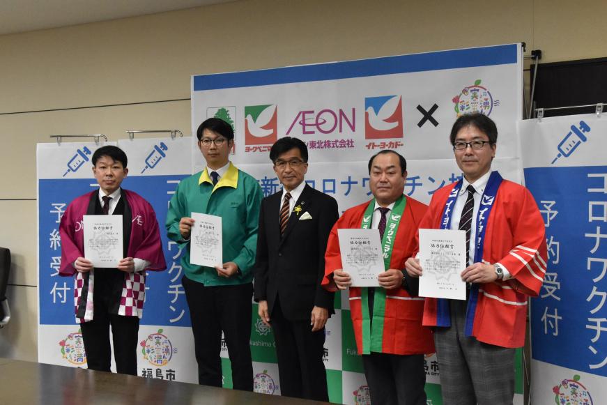 「コロナワクチン予約受付」と書かれたのぼりの前で市長の両脇に法被などを着た男性が2人ずつ立って、それぞれ協定書を持っている写真
