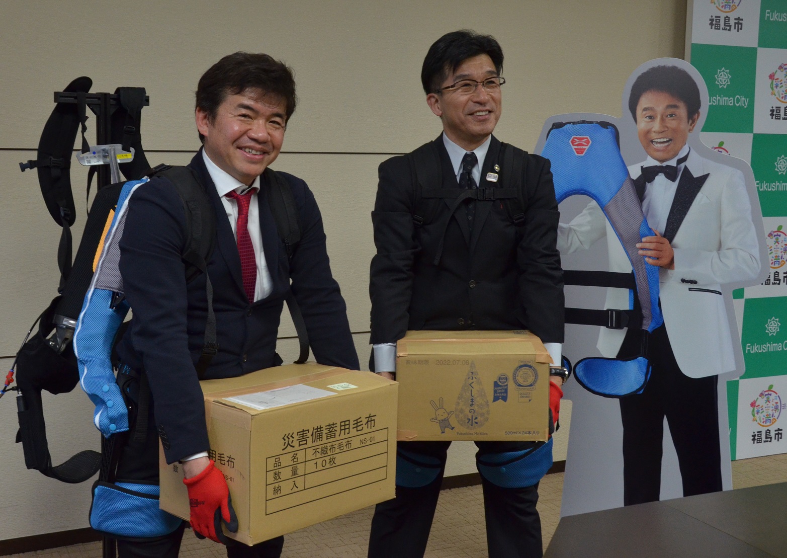 福島市のバックヤードの前にマッスルスーツを持ったダウンタウン浜田雅功さんのパネルが置かれ、その横で市長と男性が赤い手袋し、段ボール箱を持ち上げている写真