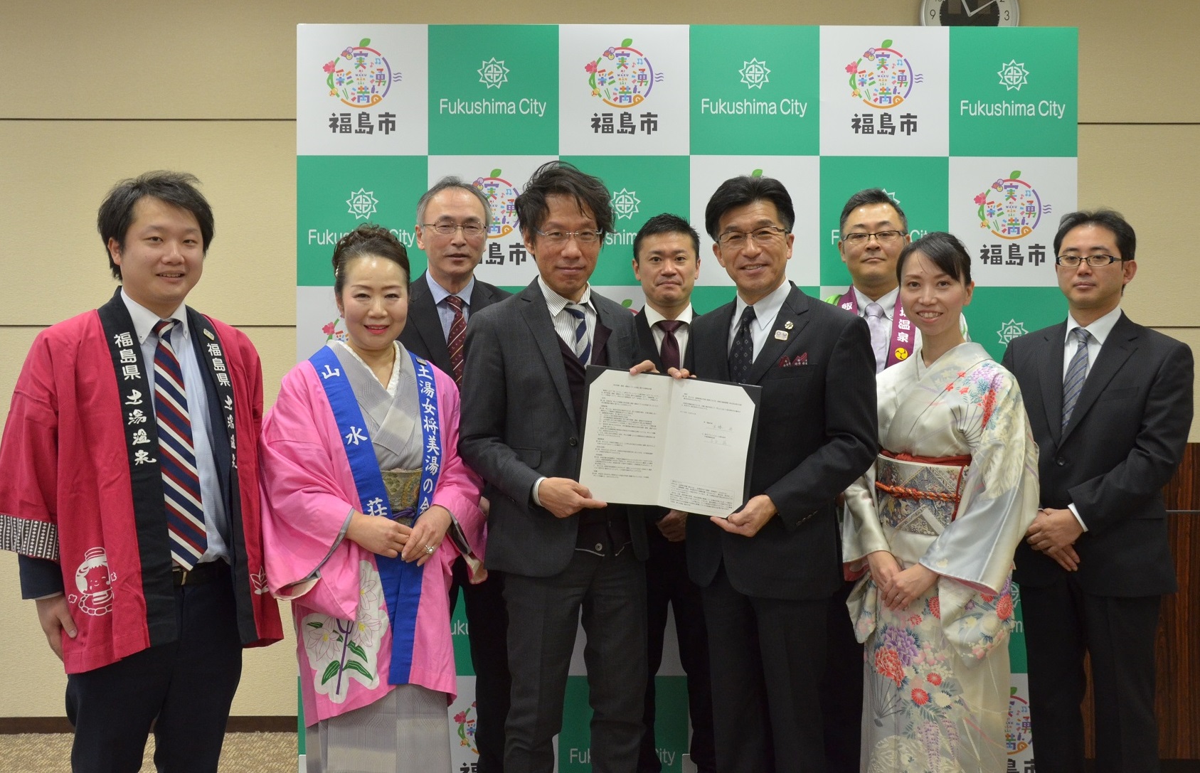 福島市のバックボードの前で2列に並び、後列に男性3名と前方に市長と男性が協定書を一緒に持ち、その両隣りに着物をきた女性と男性が並び記念撮影している写真