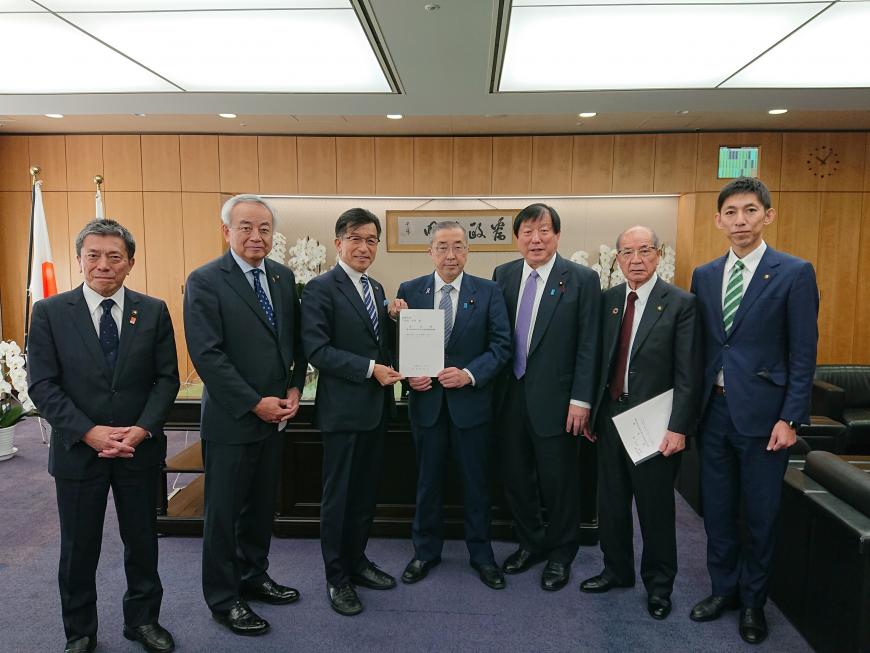 執務室内で男性7名が横に整列し、その中心で市長と男性で提言書を持っている写真