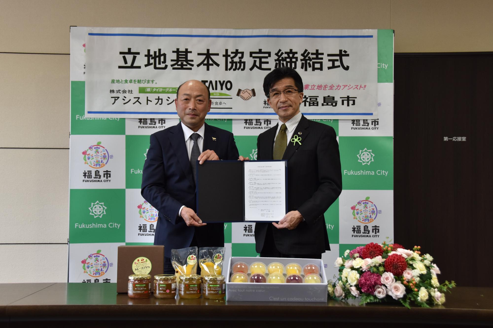 立地基本協定締結式の横断幕が貼ってあるバックパネルの前で、市長と男性で締結書ファイルを持ち、その前の机に、瓶詰めの商品、箱詰めの商品、花束が置かれている写真