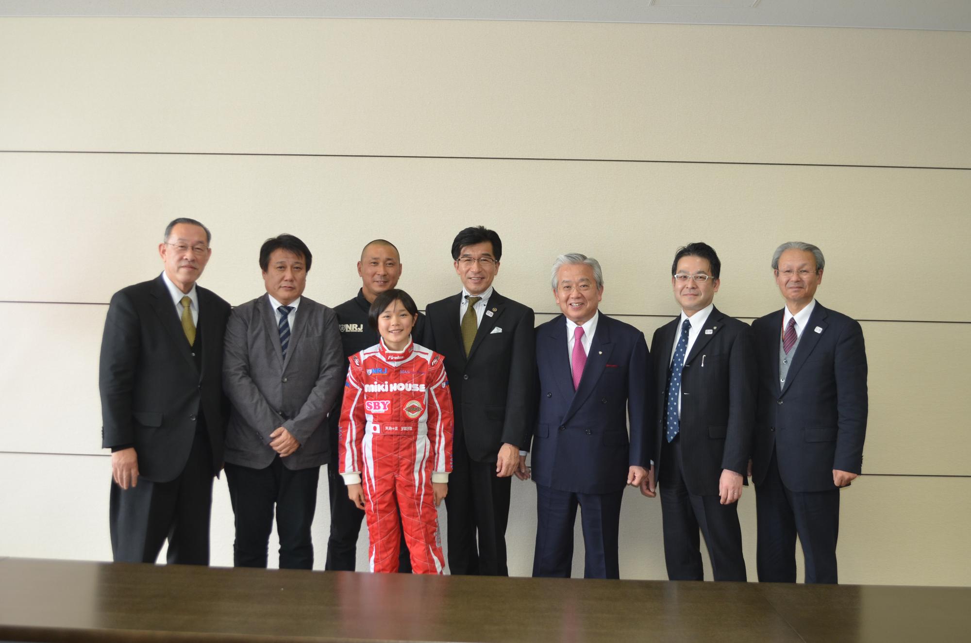 室内の壁をバックに市長の他に6名の男性が立ち、その前に赤いレーシングスーツを着た野田樹潤さんが立っている写真