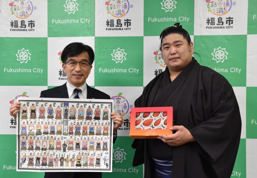 バックパネルの前で、若元春がいちごの入った箱を持ち、市長が力士が描かれた番付表を持っている写真