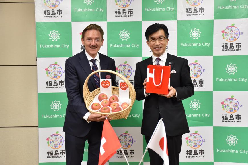 バックパネルの前で、りんごが入ったかごを持っている駐日大使と紙袋を持った市長が立っている写真