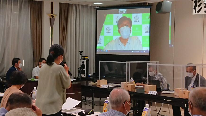 出席者たちが席に座る中、マイクを持って立った女性が、市長が映った前方の大きなモニターの方を向いて話をしている写真