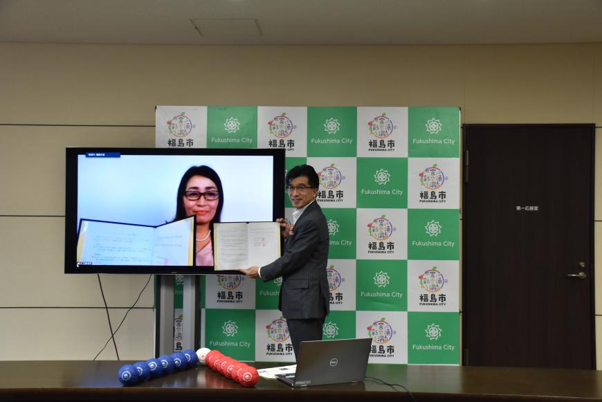 福島市のバックボードの前に女性が映ったモニターが置かれ、市長が協定書も持って横に並び記念撮影している写真