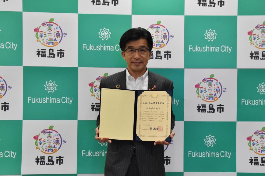 福島市のバックボードの前で市長がSDGz認定選定証を持った写真