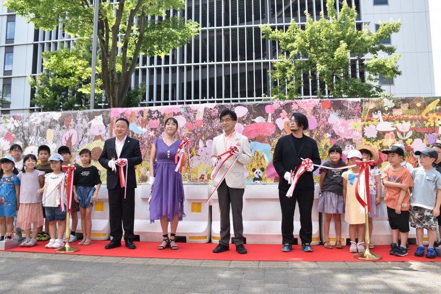 建物と植え込みの木の前にカラフルな絵が描かれたフェンスが設置されており、その前に敷かれた赤いカーペットに並んで立った子供たちの間に立った市長、2名の男性、紫のドレスの女性がテープカットをしている様子の写真