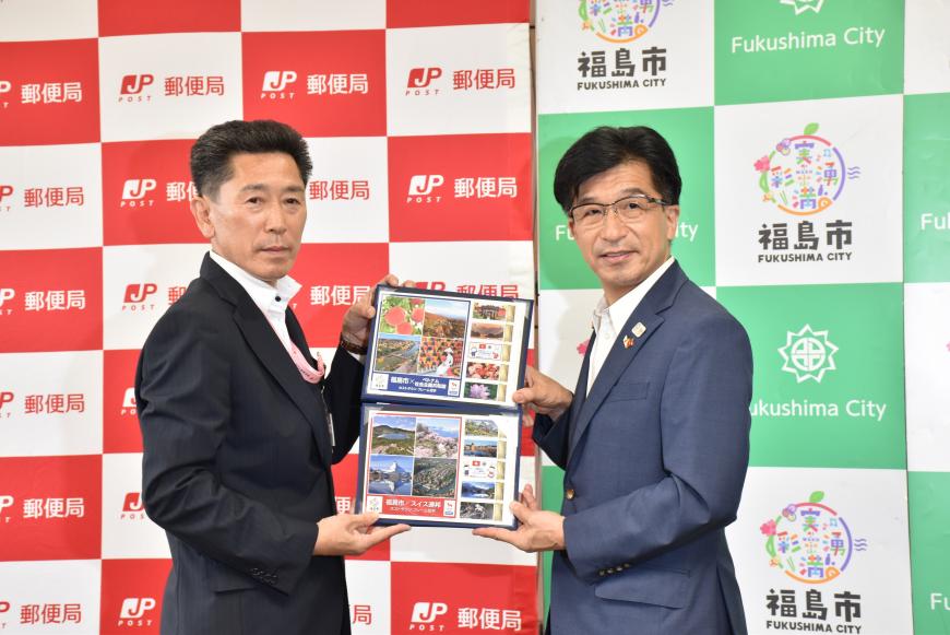 郵便局と福島市のバックボードが並べて置かれた前で市長と日本郵便の男性が両側から切手のつづりを持って写った記念写真