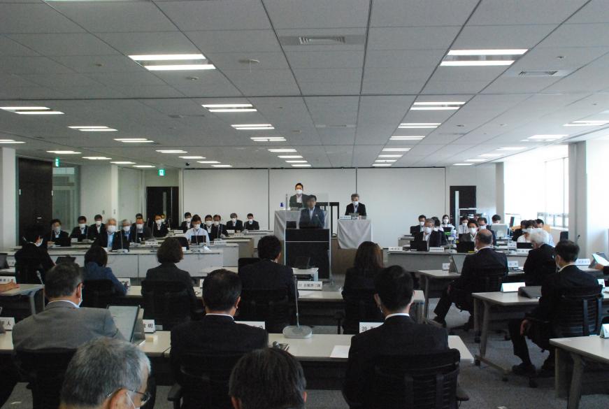 大勢の出席者たちが集まった市議会の会場で、アクリルボードに囲まれた演台に立った市長が話をしている様子の写真