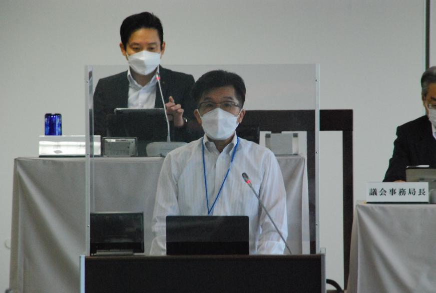 後方から男性2名が見ている前で、市長が演台に立ち、話をしている写真