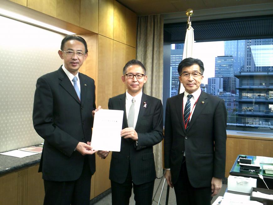 執務室内で市長の横の男性2名で書類を持ち並んでいる写真