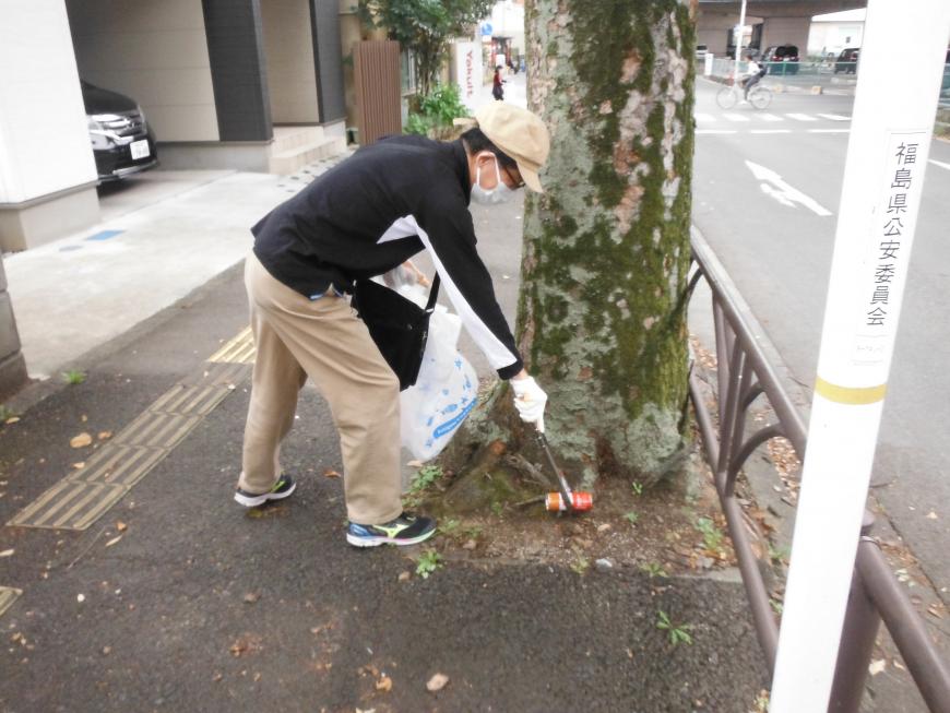 ゴミ収集のビニール袋を持った人が木の根元に落ちている空き缶を火ばさみで掴んでいる写真