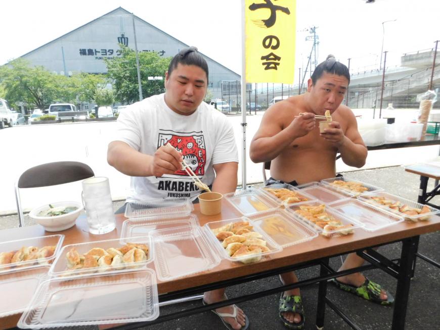 力士2名が椅子に座り、パックに入って机に並べられた餃子を食べている写真