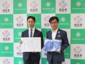バックパネルの前で、市長が青の荷物、隣の男性が賞状を持ち立っている写真