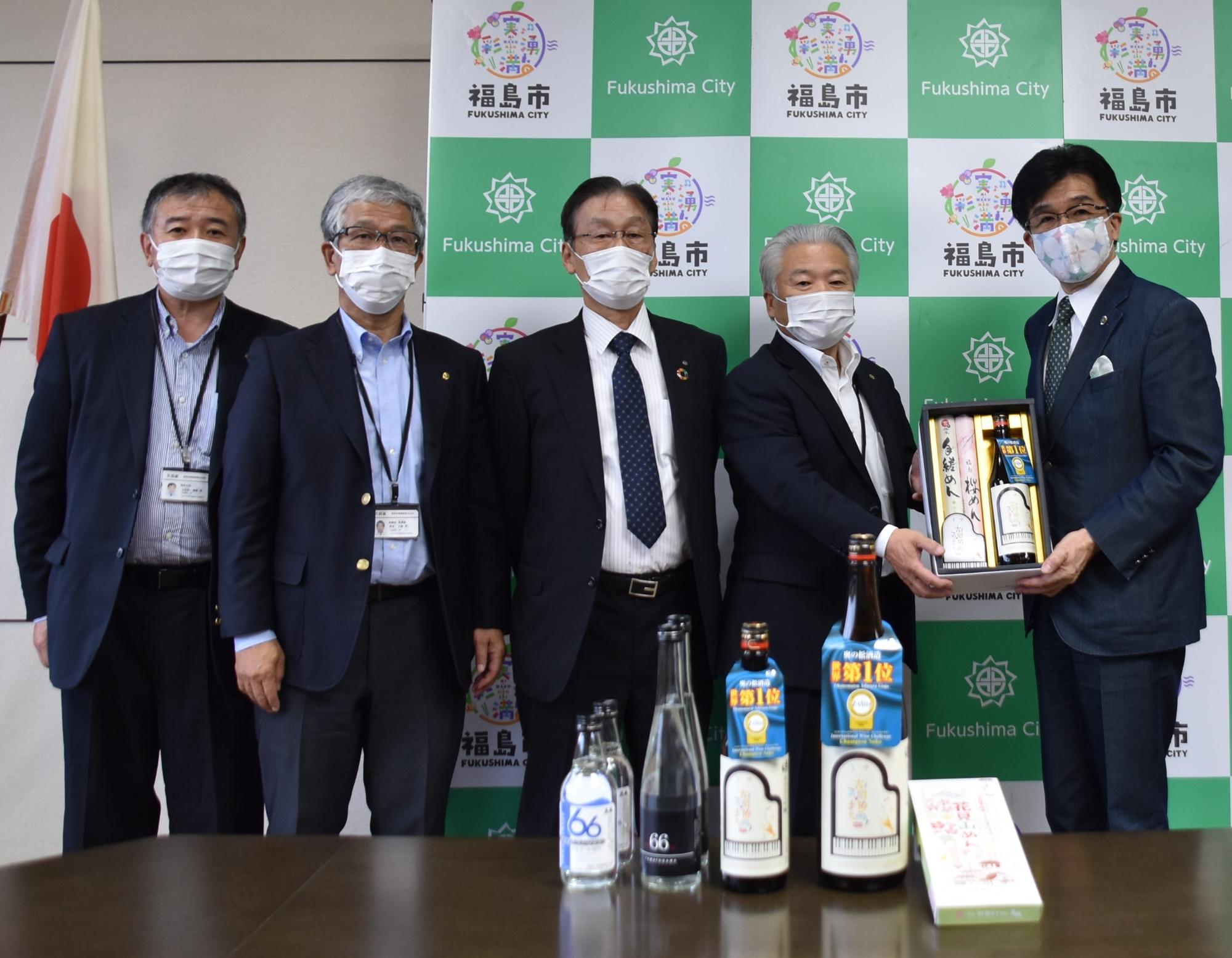机の上にはお酒が並べられており、福島市のバックボードの前で4名の男性が並び、そのうちの一人と市長がお酒の入った箱を一緒に持って記念撮影している写真