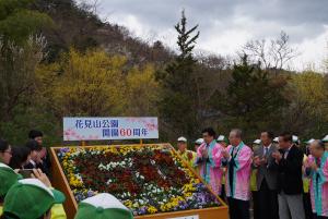 「花見山公園開園60周年」と書かれた看板と大きな木箱に黄色や紫色の花で囲まれ濃い赤色の花で60の文字が作られた花アートの周りで、大勢の人たちが拍手をしている除幕式の様子の写真