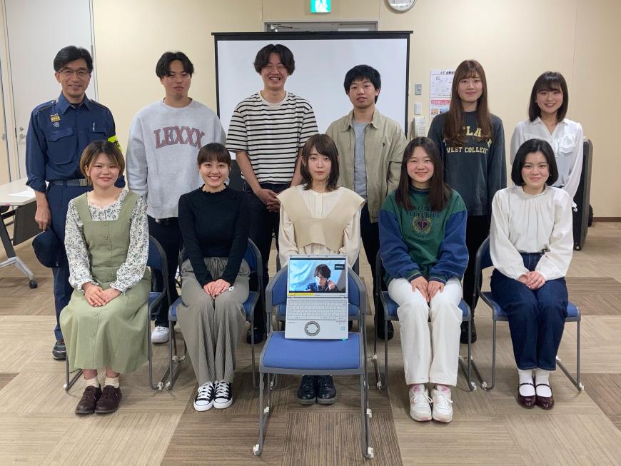 手前に置いた椅子に男性が映ったパソコンが置かれ、その後ろに設置された椅子に5名の女性が座り、後方に市長と男女5名が立ち記念撮影している写真