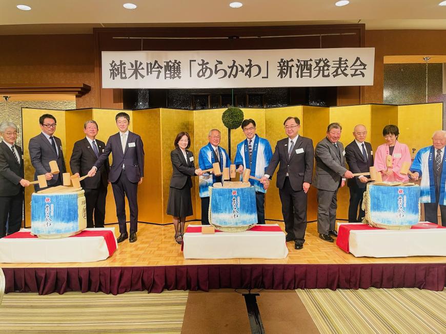 金屛風の前の3個の酒樽にそれぞれ4名が立ち、鏡開きをしている写真