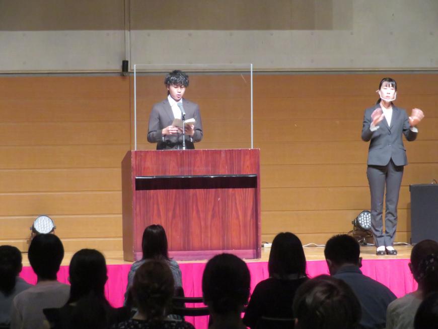 多くの参加者の前で、演台で男性が話をしている横で女性が手話通訳をしている写真
