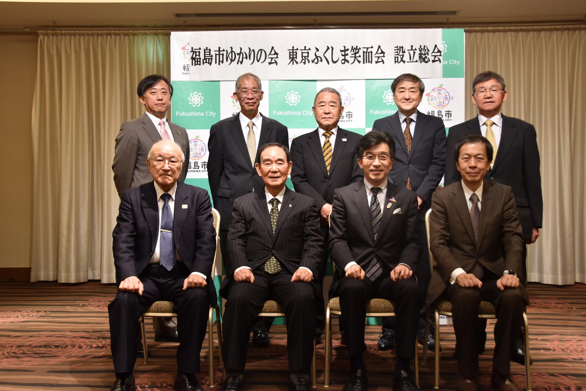 福島市ゆかりの会東京ふくしま笑而会設立総会と書かれた横断幕が貼られたバックパネルの前で、市長を含む男性4人が座り、後列に男性5人が立って撮影した写真
