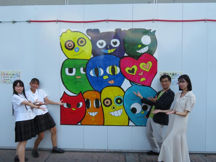 白地の仮囲いに描かれた作品の両脇で生徒2名と市長と女性が両手を作品に向けている写真