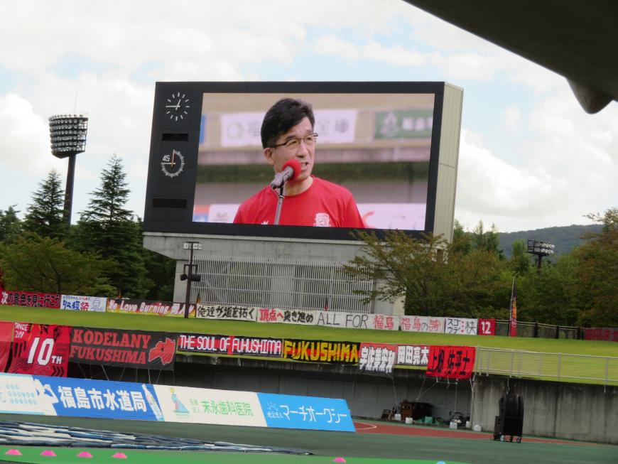 横断幕が掛けられているスタジアムの大型ビジョンに赤のユニフォーム姿の市長が映し出されている写真