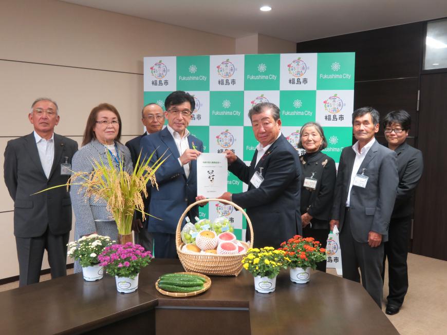 果物が入ったかご、稲穂、鉢植えの花が置かれた机を前にして、農業委員の男性4人、女性2人の中心で市長と委員の代表の男性で意見書を持っている写真