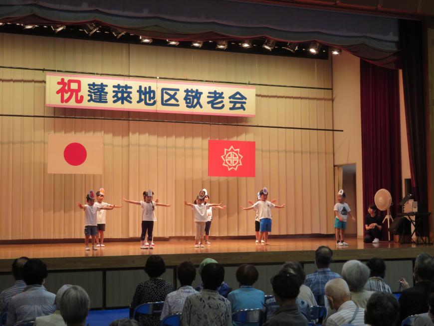 「蓬莱地区敬老会」と書かれた横断幕があるステージで、ご年配の方が観客席で見ている中、園児たちが両手を広げて踊りをしている写真