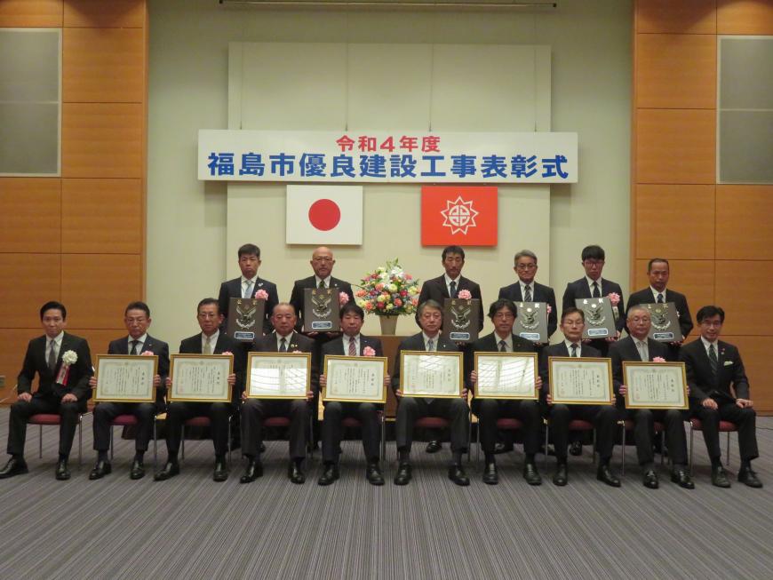 表彰式で前列に額入りの賞状を持った男性8名と市長が座り、後列に盾を持った男性6名が立って撮影した写真