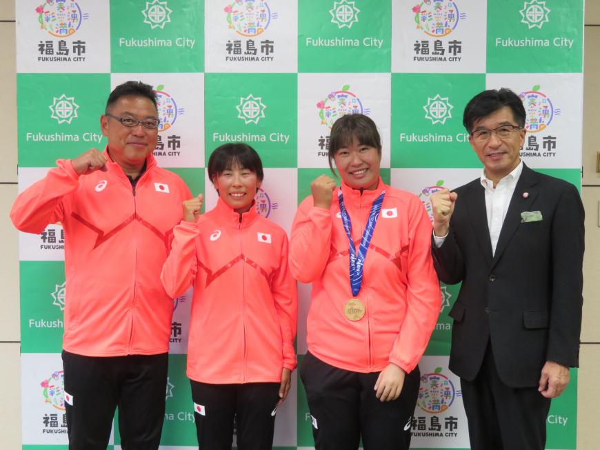 バックパネルの前で、市長と日本代表のジャージを着た女性2名のうち1名がメダルをかけて、他に男性1名が立ち、右手でガッツポーズをしている写真
