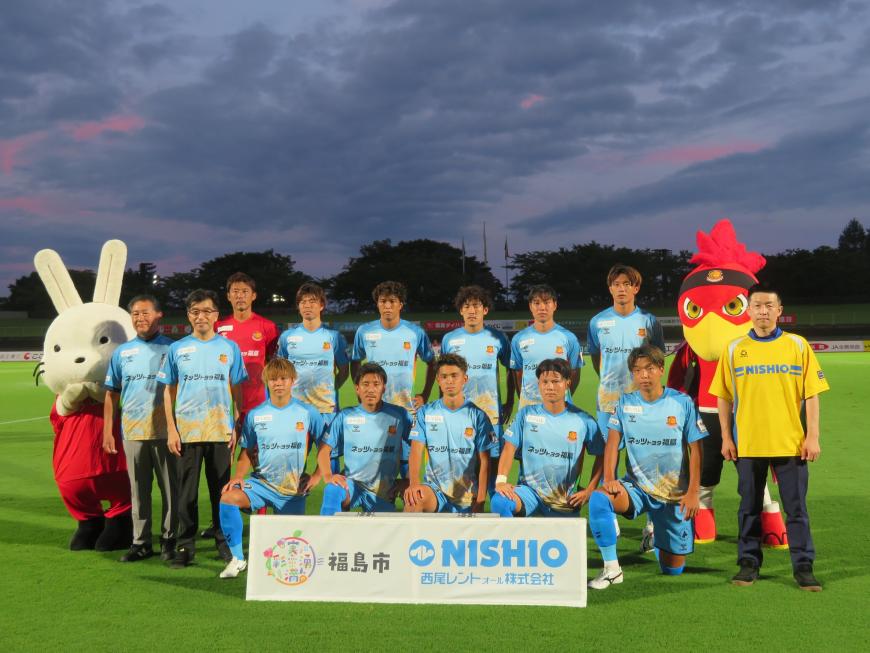 グランドでサッカーチームの選手と市長と関係者の方と2体のマスコットキャラクターが並んでいる集合写真