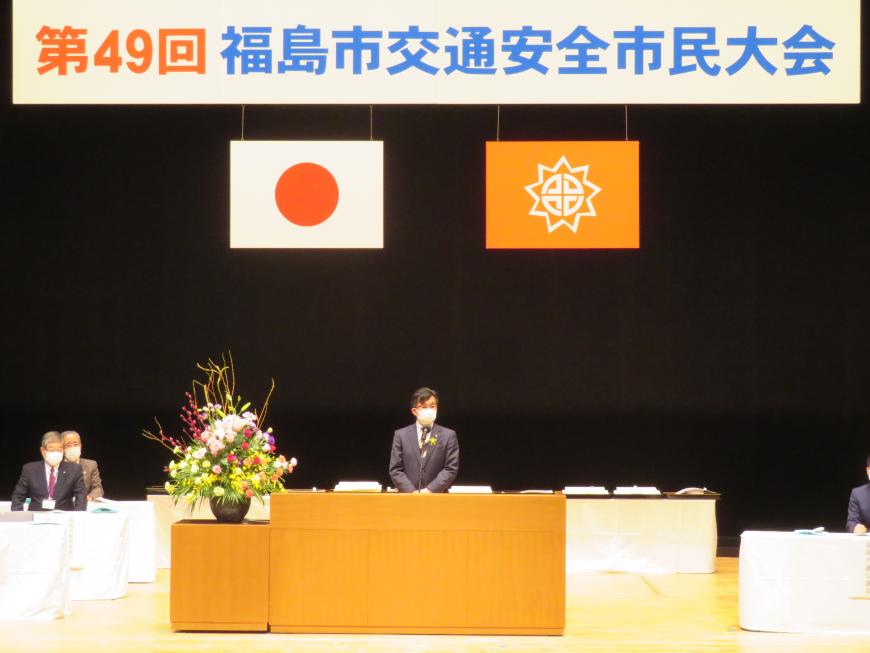 福島市交通安全市民大会の横断幕がある会場で、他の来賓の方々が座っているステージで市長が演台に立って話をしている写真