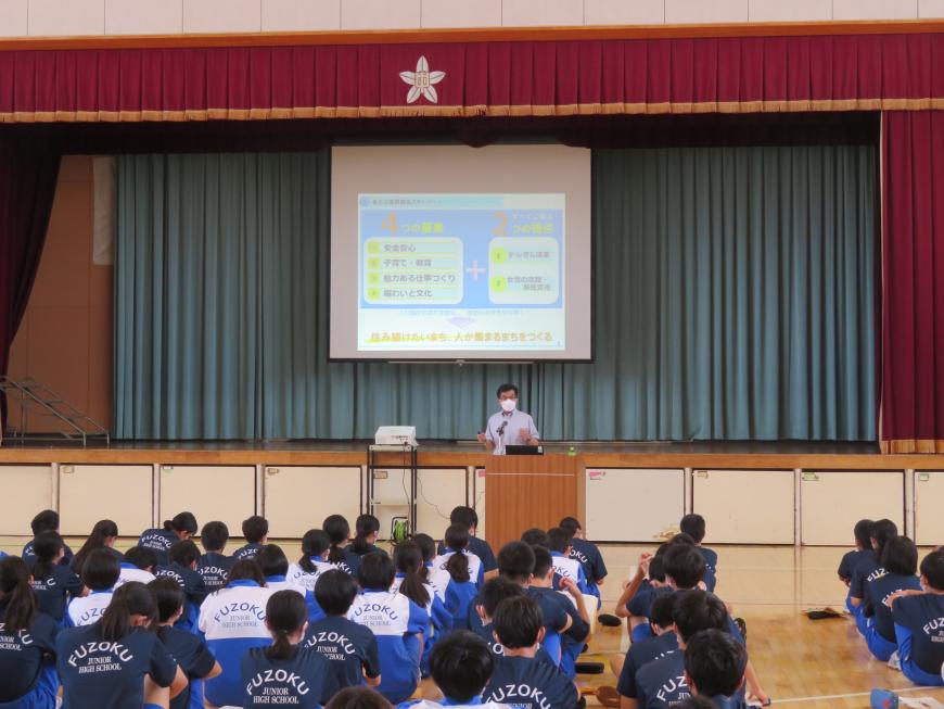 体育館で座っている生徒の前で、市長がステージのスクリーンに映し出された画像をバックに話をしている写真