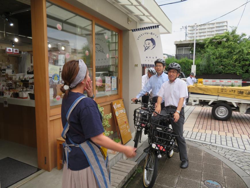 市長と男性がヘルメットを被り、自転車に乗った状態で店頭で女性と話をしている写真