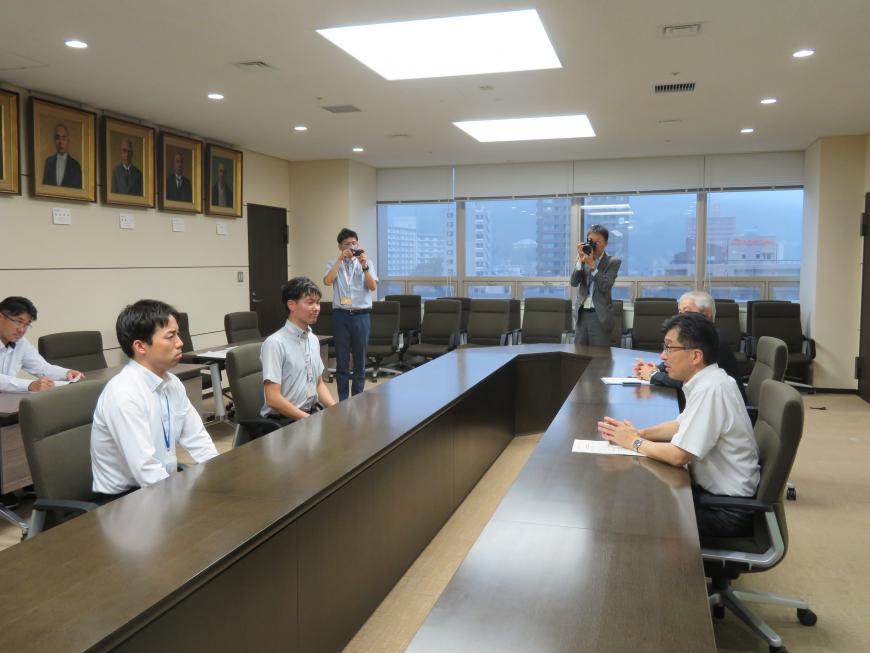 執務室内で楕円形状の長机に座り、職員2名に向かって市長が話をしている写真