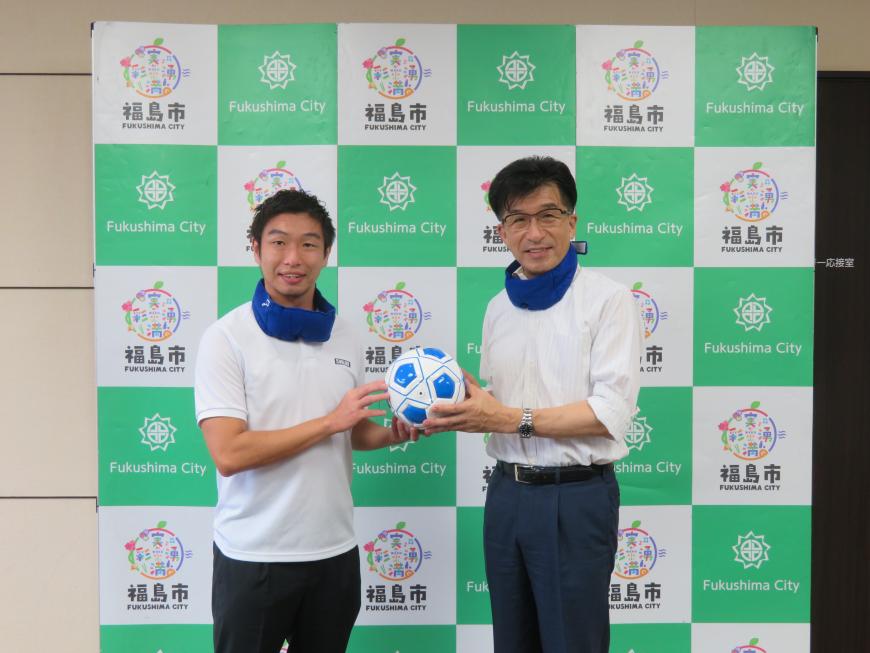 バックパネルの前で、市長と男性が首にアイマスクを巻いて、サッカーボールを持っている写真