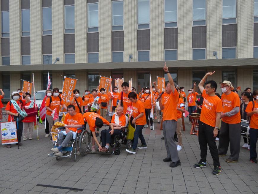 広場に車いすの方、市長をはじめ多くのサポートする方がお揃いのオレンジ色のTシャツを着て集まっている写真