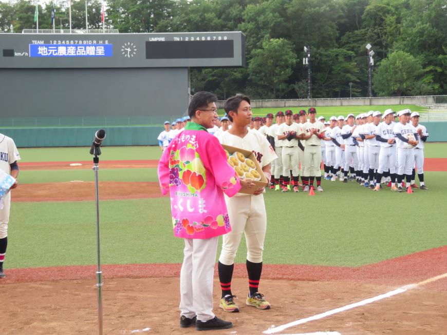 野球場で法被を着た市長が出場する選手の代表に果物が入った段ボールを渡している写真