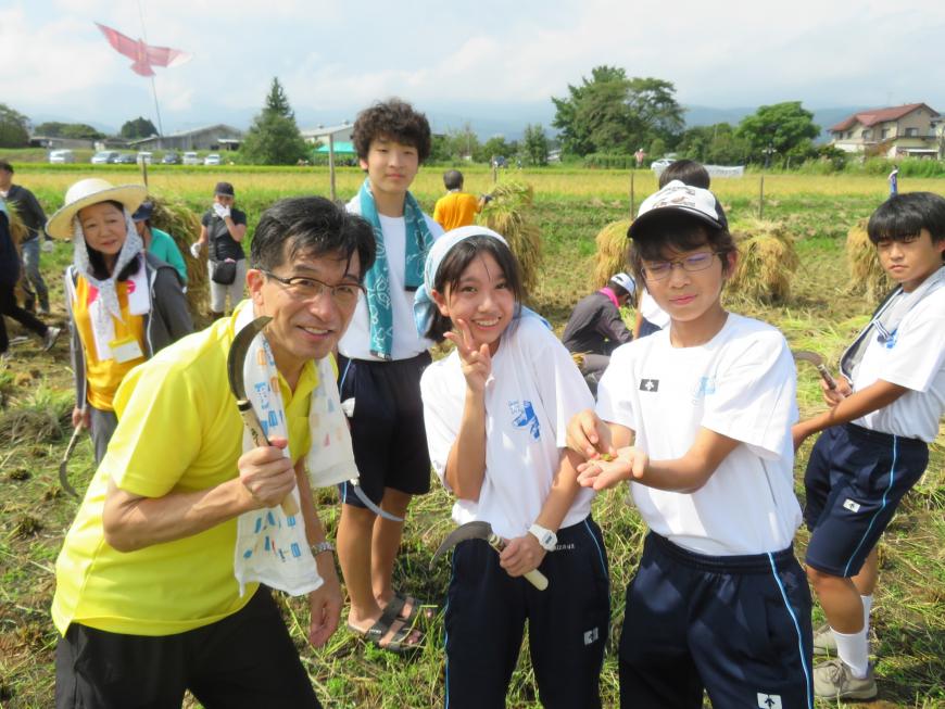 田んぼで稲刈りを行立ている最中に、市長と生徒が鎌を手にしてカメラに向かってポーズを決めている写真