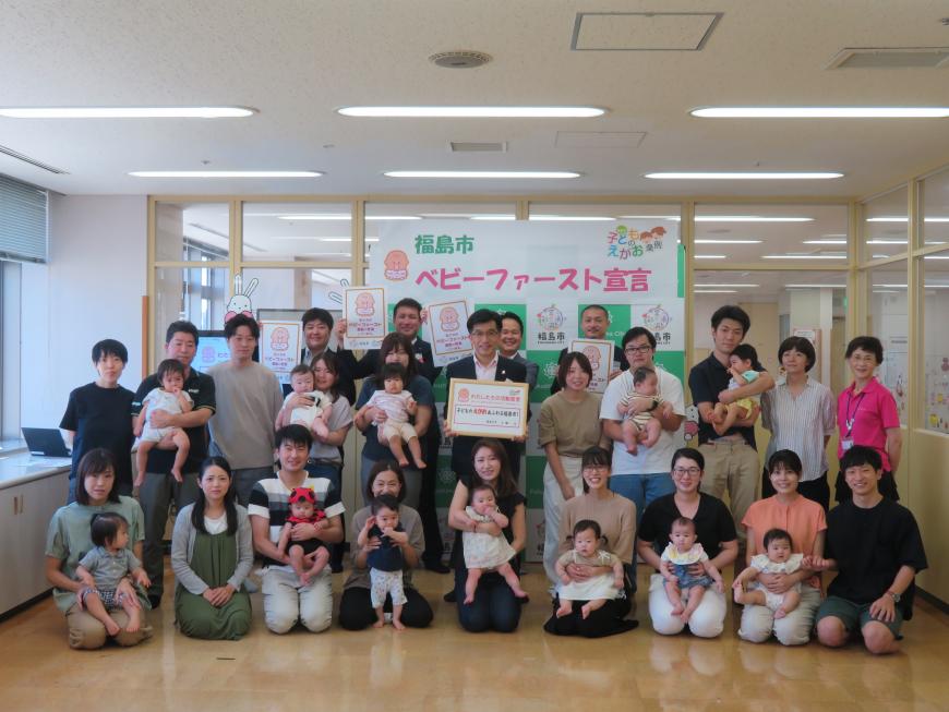 フローリングの部屋で市長を中心にして赤ちゃんを抱っこした保護者の皆さんが集合して撮影した写真