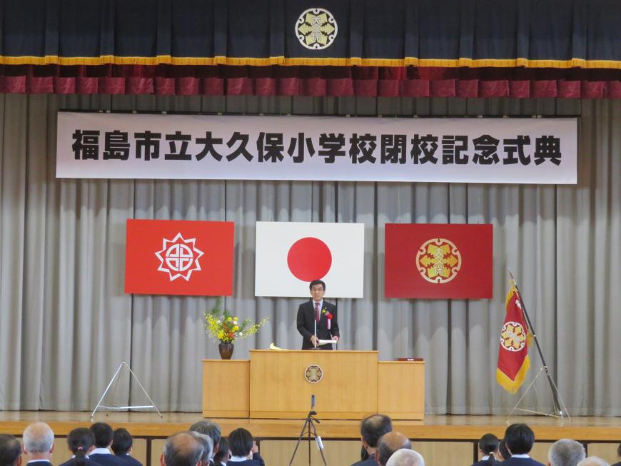 大久保小学校閉校記念式典の横断幕がある体育館で、参加者の前で市長がステージの演台に立って話をしている写真