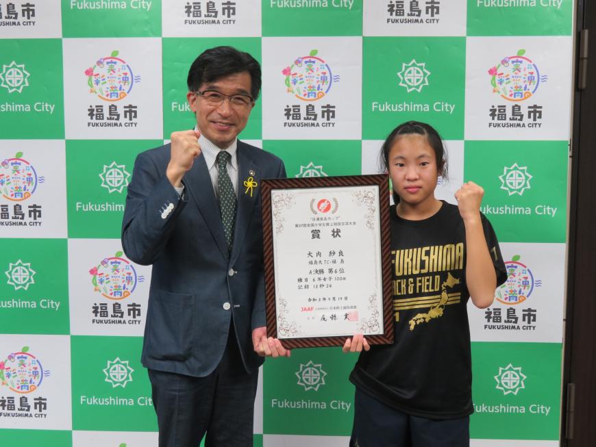 バックパネルの前で、市長と女子児童で額入りの賞状を持ってガッツポーズをしている写真