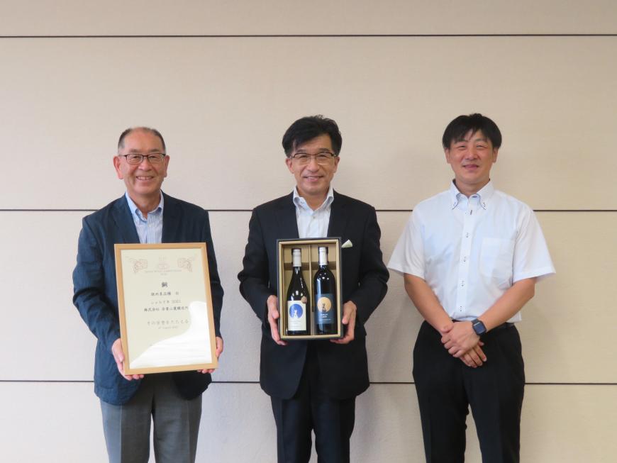 額入りの賞状を持った男性と半袖シャツの男性の間に市長がワインボトルが2本入ったケースを持って立っている写真