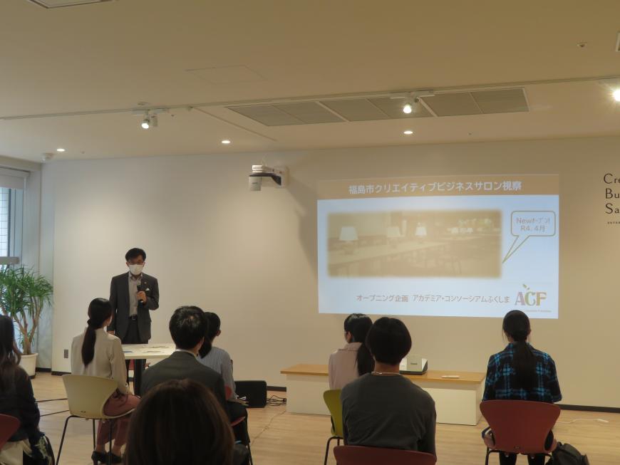 参加者が座ったいる前に映し出された映像の横で、市長が説明をしている写真