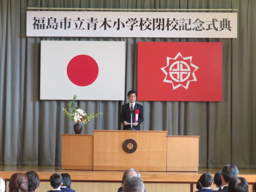 青木小学校閉校記念式典の横断幕が吊るされた体育館で、参加者の前で市長がステージの演台に立って話をしている写真