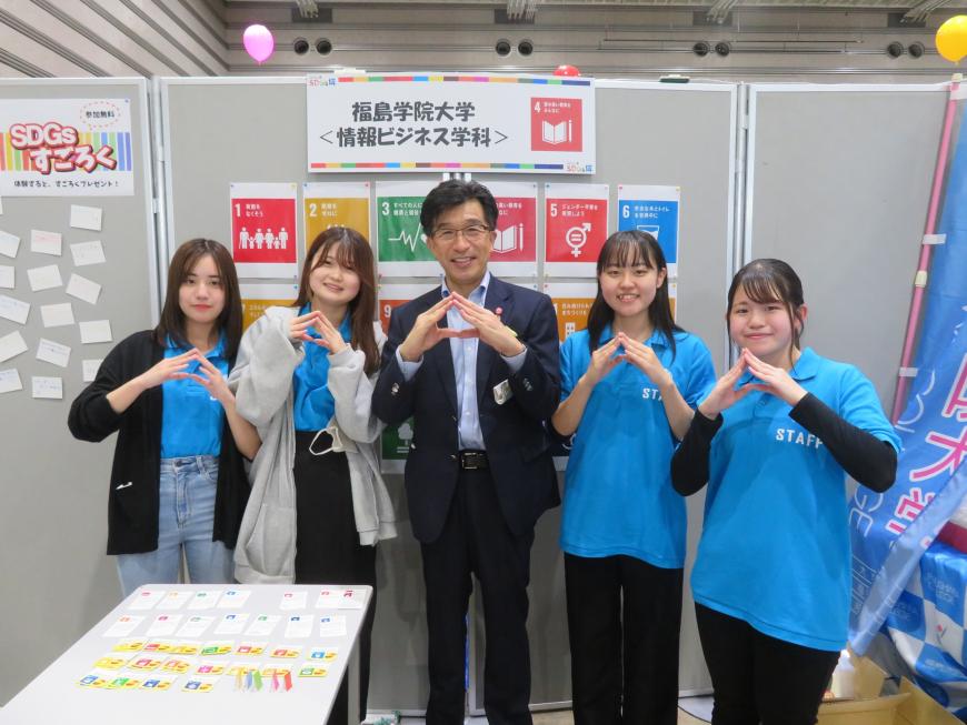 福島学院大学情報ビジネス科のブースで女子学生4名の中心に市長が立ち、皆で両手の指先を合わせたポーズをしている写真