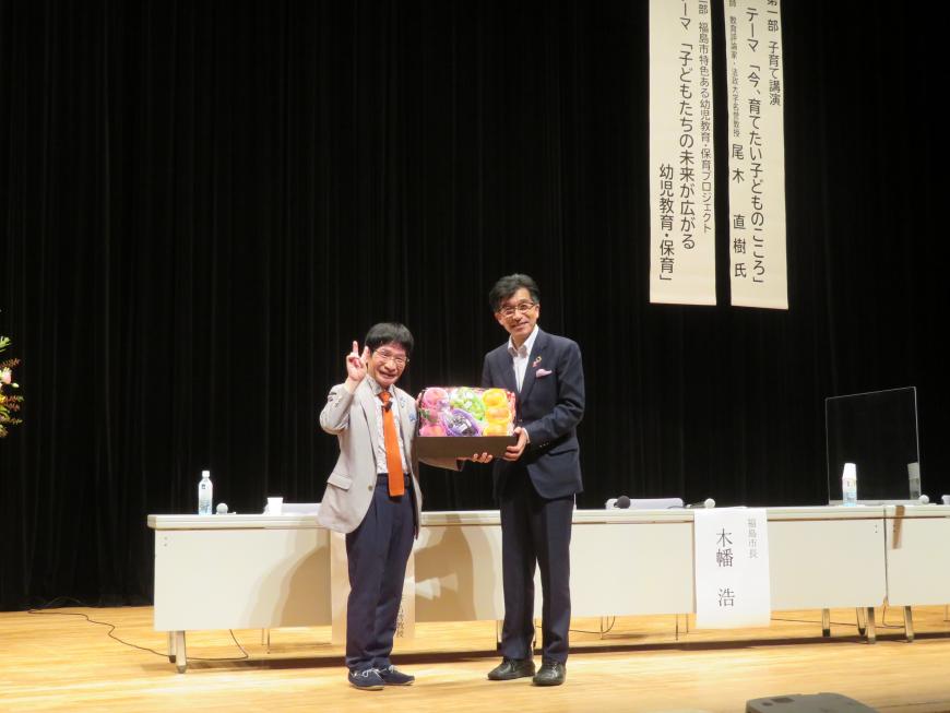 ステージで市長とピースサインをしている尾木ママでフルーツが入った箱を持っている写真