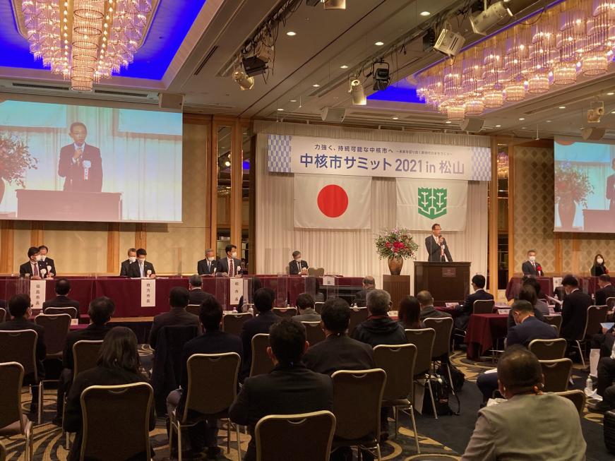 中核市サミットの会場で演台に立って話をしている男性の姿が両脇にあるスクリーンに映し出されている写真